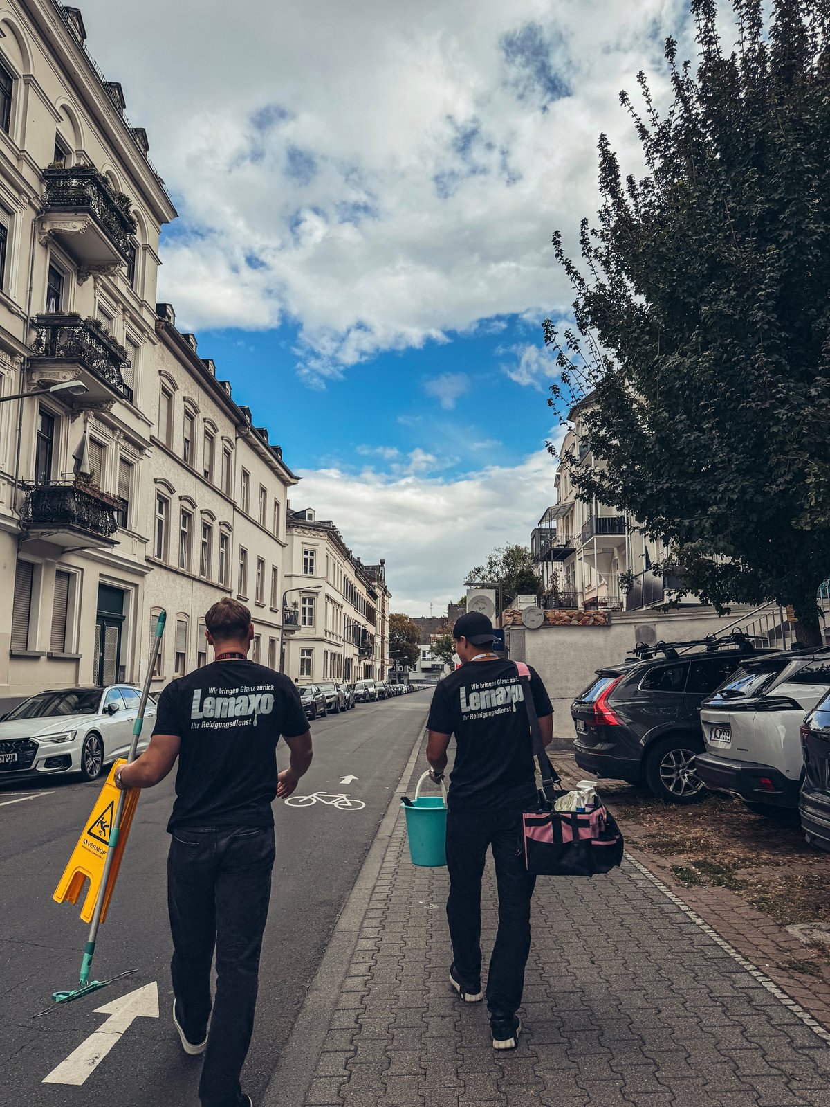 Two workers in Lemaxo shirts walking down an urban European street lined with elegant buildings and cars, carrying cleaning equipment